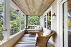 A covered porch with a sofa and wicker chairs, large windows, and a view of trees. - 3BR lakefront home: pvt beach, dock, gazebo (Center Harbor)