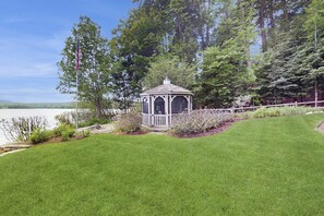 A gazebo with a white railing, surrounded by a well-maintained lawn and a variety of shrubs. - 3BR lakefront home: pvt beach, dock, gazebo (Center Harbor)