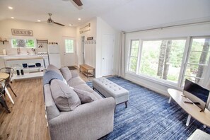 A modern living room with a sectional sofa, a television, and a large window offering a view of trees. - 1BR cottage with beach access & dock (Alton)