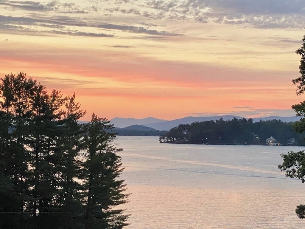 A calm lake with a sunset sky, surrounded by trees and distant mountains.