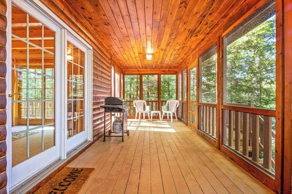 A wooden porch with a grill and chairs, large windows, and a welcome mat.