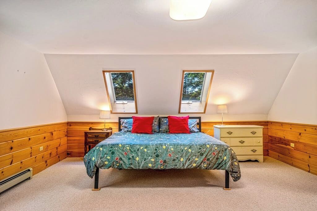 Une chambre avec un plafond en pente, un lit recouvert d’une couvre-lit à motifs floraux, deux lampes de chevet, une commode et deux fenêtres.