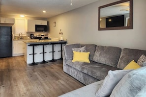 A modern kitchen with a central island, bar stools, and a grey sofa with yellow cushions. - 1BR with pool & tennis, walk to Lake Winnipesaukee (Gilford)