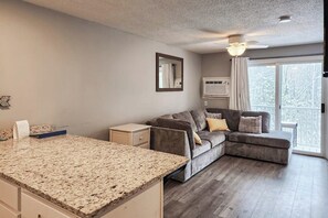A modern kitchen with a granite countertop, a grey sectional sofa with cushions, a small white nightstand, and a window with curtains. - 1BR with pool & tennis, walk to Lake Winnipesaukee (Gilford)