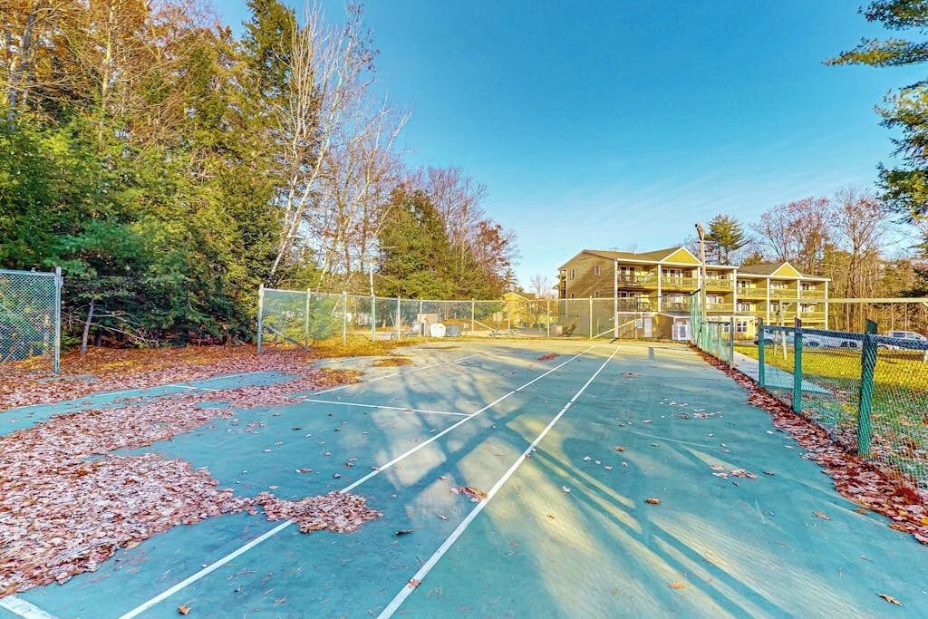 An outdoor tennis court with a net, surrounded by trees and a building in the background.