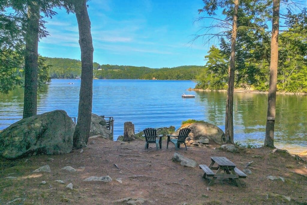 A lakeside campsite with a picnic table and chairs, surrounded by trees and a clear view of the lake.