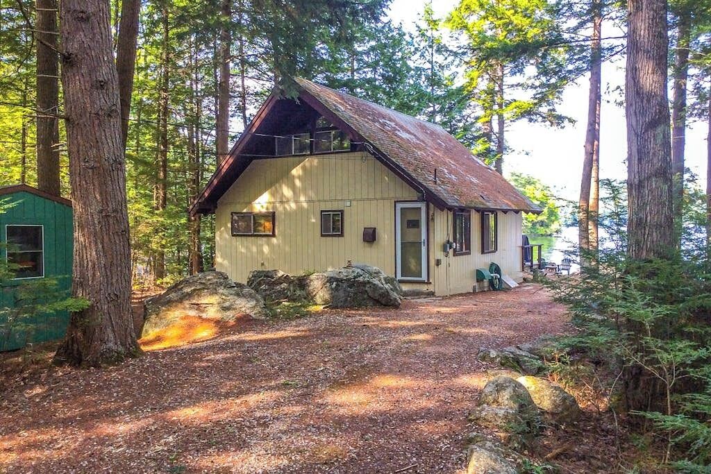 Un chalet rustique avec un toit en pente, entouré de grands arbres et bordé d’une allée en gravier.
