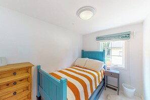 A bedroom with a single bed, a wooden dresser, a nightstand, a window with a curtain, and a white trash bin. - 4BR lakeside cottage with dock access, mtn views (Meredith)