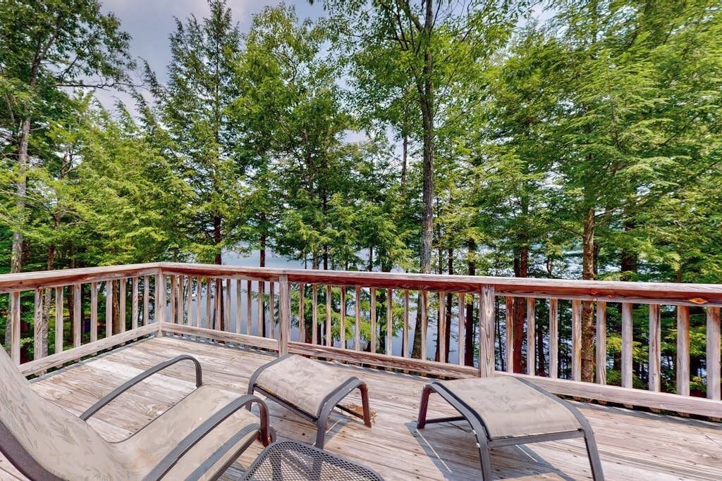 Une terrasse en bois avec des chaises en métal, donnant sur un lac entouré d’arbres.