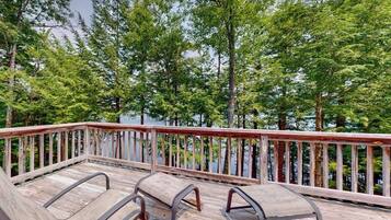 Une terrasse en bois avec des chaises en métal, donnant sur un lac entouré d’arbres.
