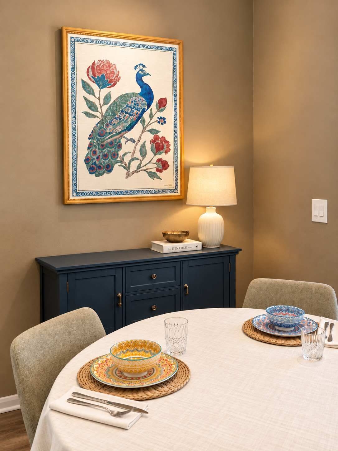 Een eetgedeelte met een ronde tafel gedekt voor twee personen, een schilderij van een pauw in een lijst erboven, een donkerblauwe kast eronder en een witte lamp aan de rechterkant.