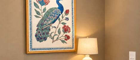 Een eetgedeelte met een ronde tafel gedekt voor twee personen, een schilderij van een pauw in een lijst erboven, een donkerblauwe kast eronder en een witte lamp aan de rechterkant.