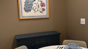 Un área de comedor con una mesa dispuesta para dos, una obra de arte enmarcada de un pavo real y un gabinete negro debajo.