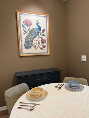 A dining area with a table set for two, a framed artwork of a peacock, and a black cabinet below. - The Nomad / Domain Rolex Building (Austin)