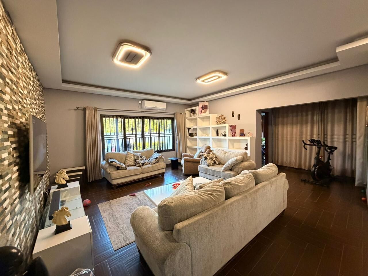 A spacious living room with a large sofa, a coffee table, and a television. There is a bookshelf with decorative items and a window with curtains.