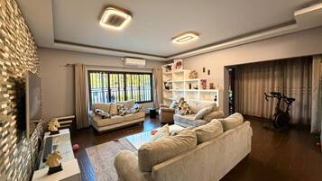 A spacious living room with a large sofa, a coffee table, and a television. There is a bookshelf with decorative items and a window with curtains.