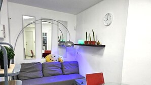 Una sala de estar con sofá, una mesa de vidrio y una silla roja. Hay un reloj en la pared y una estantería con plantas.