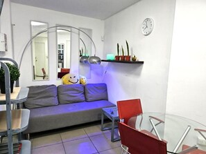 Una sala de estar con sofá, una mesa de vidrio y una silla roja. Hay un reloj en la pared y una estantería con plantas.