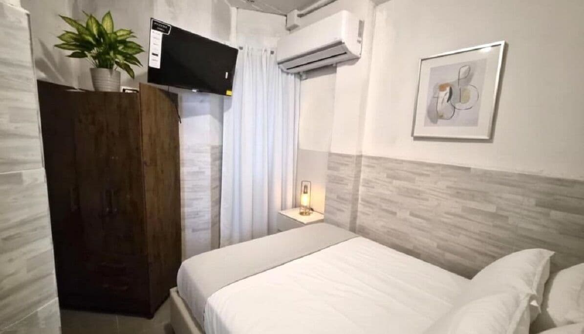 A hotel room with a bed, a TV mounted on the wall, an air conditioning unit, a potted plant, a framed artwork, and a bedside table with a lamp.