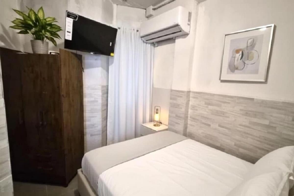 A hotel room with a bed, a TV mounted on the wall, an air conditioning unit, a potted plant, a framed artwork, and a bedside table with a lamp.