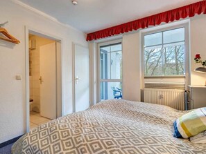 A bedroom with a patterned bedspread, a radiator, a window with a view, and a door leading to a bathroom. - Residenz am Buchenpark App. 14 (Heringsdorf)