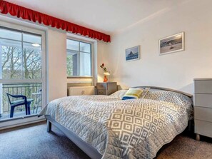 Ein Schlafzimmer mit Bett, Kommode, Heizkörper, Fenster mit Blick auf Bäume und einem Balkon mit einem Stuhl.