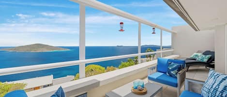 A balcony with a view of the sea, blue cushions, a small table, and a chair.
