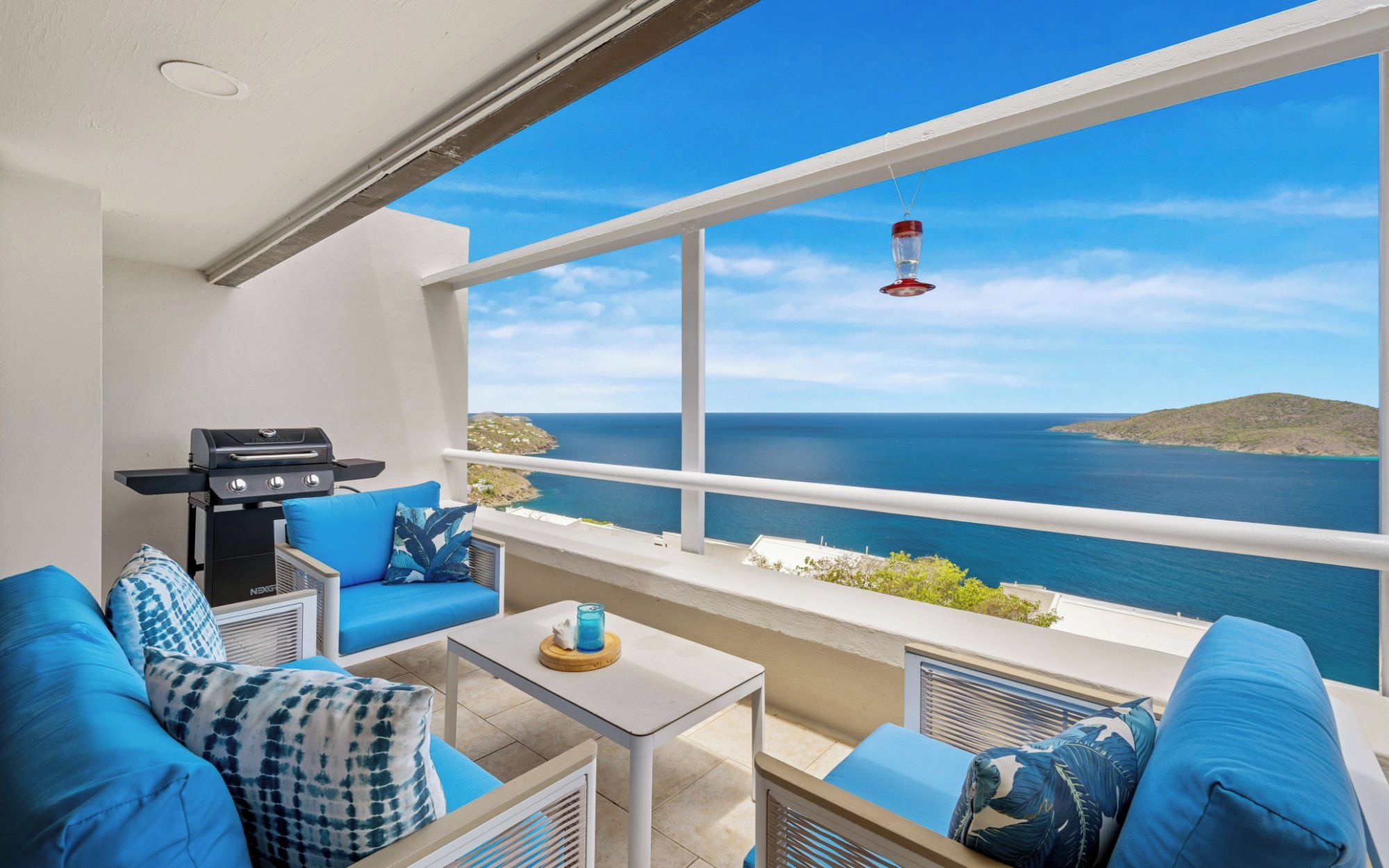 Un balcón con vista al mar, una parrilla y mobiliario tapizado en azul.