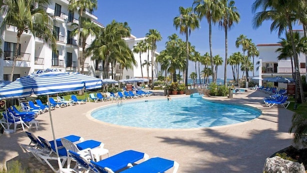 Zona de piscina con sillones de descanso y sombrillas, rodeada de palmeras y un edificio con balcones.