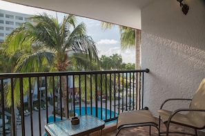 Un balcón con mesa y sillas de vidrio, con vistas a una piscina y palmeras.