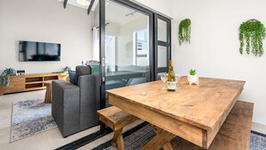 Ein modernes Wohnzimmer mit einem Holzesstisch, einem grauen Sofa, einem Fernseher und einer Glastür, die auf einen Balkon führt.