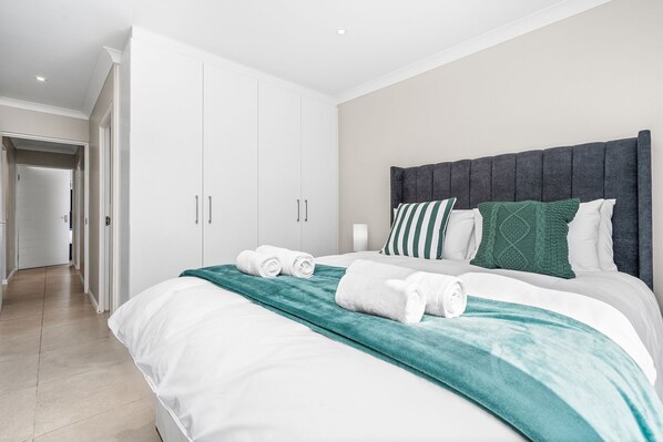 Una habitación de hotel con una cama grande, cortinas blancas, un cabecero gris y una manta turquesa.