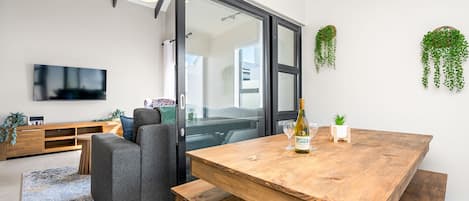 Una sala de estar moderna con una mesa de comedor de madera, un sofá gris, un televisor y un gran ventanal corredizo que da a un balcón.