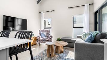 A modern living room with a flat-screen TV, a dining table with chairs, a sofa, a patterned armchair, and a coffee table.
