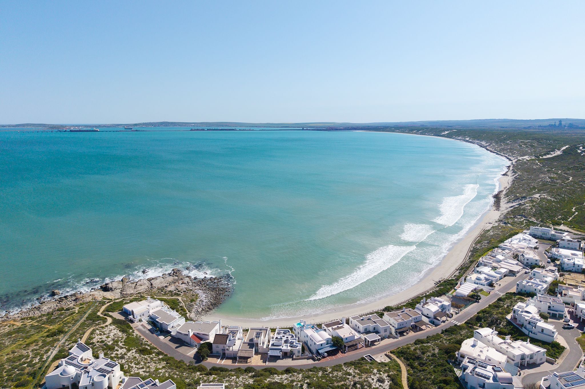 An aerial view of a coastal area with a beach, a rocky shoreline, and a cluster of white buildings with flat roofs.