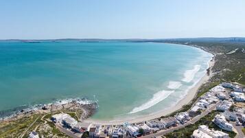 An aerial view of a coastal area with a beach, a rocky shoreline, and a cluster of white buildings with flat roofs.