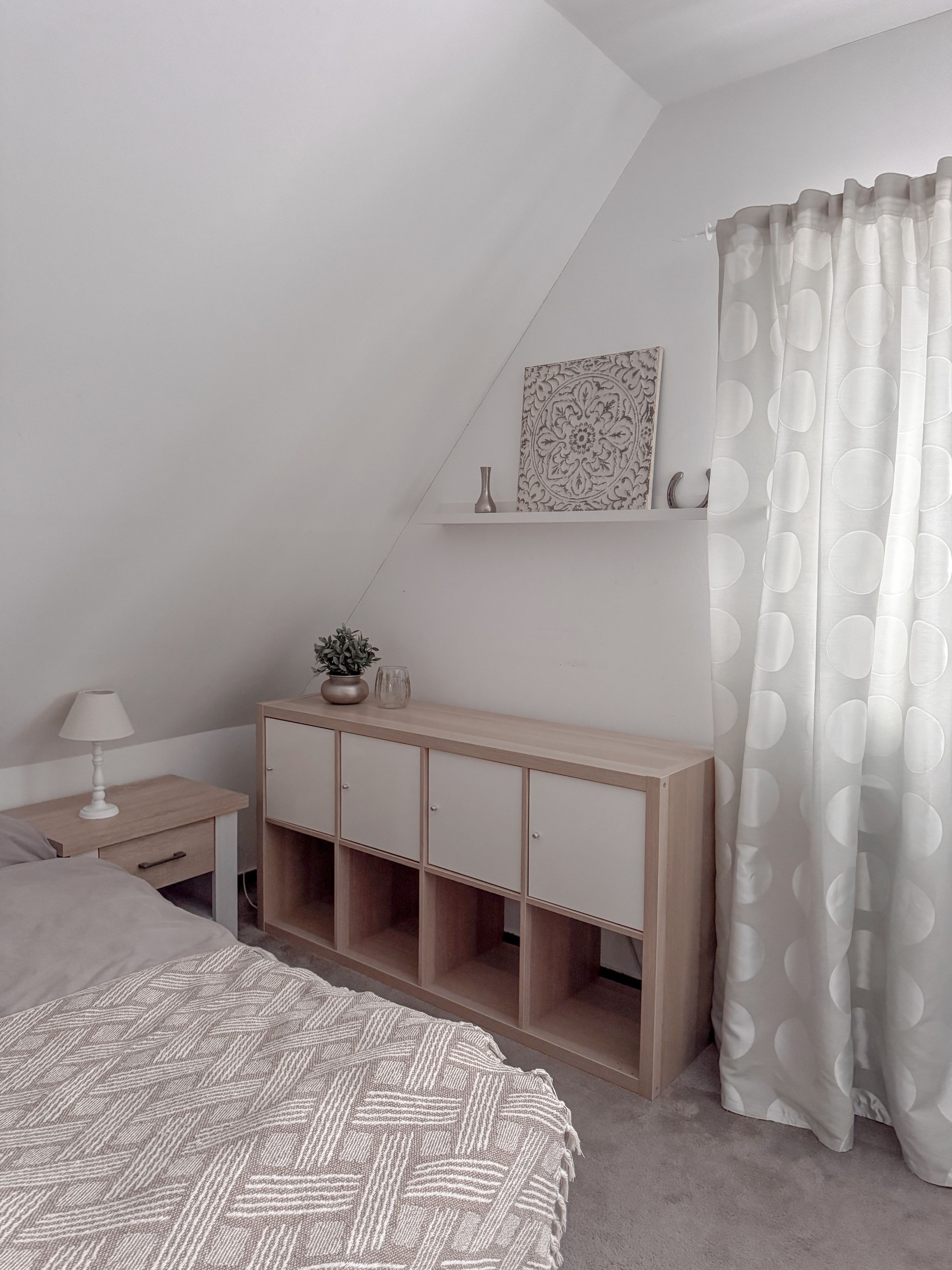 Ett sovrum med ett sluttande tak, en säng med ett mönstrat överkast, ett sängbord med en lampa, en säng med träkonsol och öppna hyllor, en vägghylla med dekorativa föremål och en gardin med ett cirkelformat mönster.