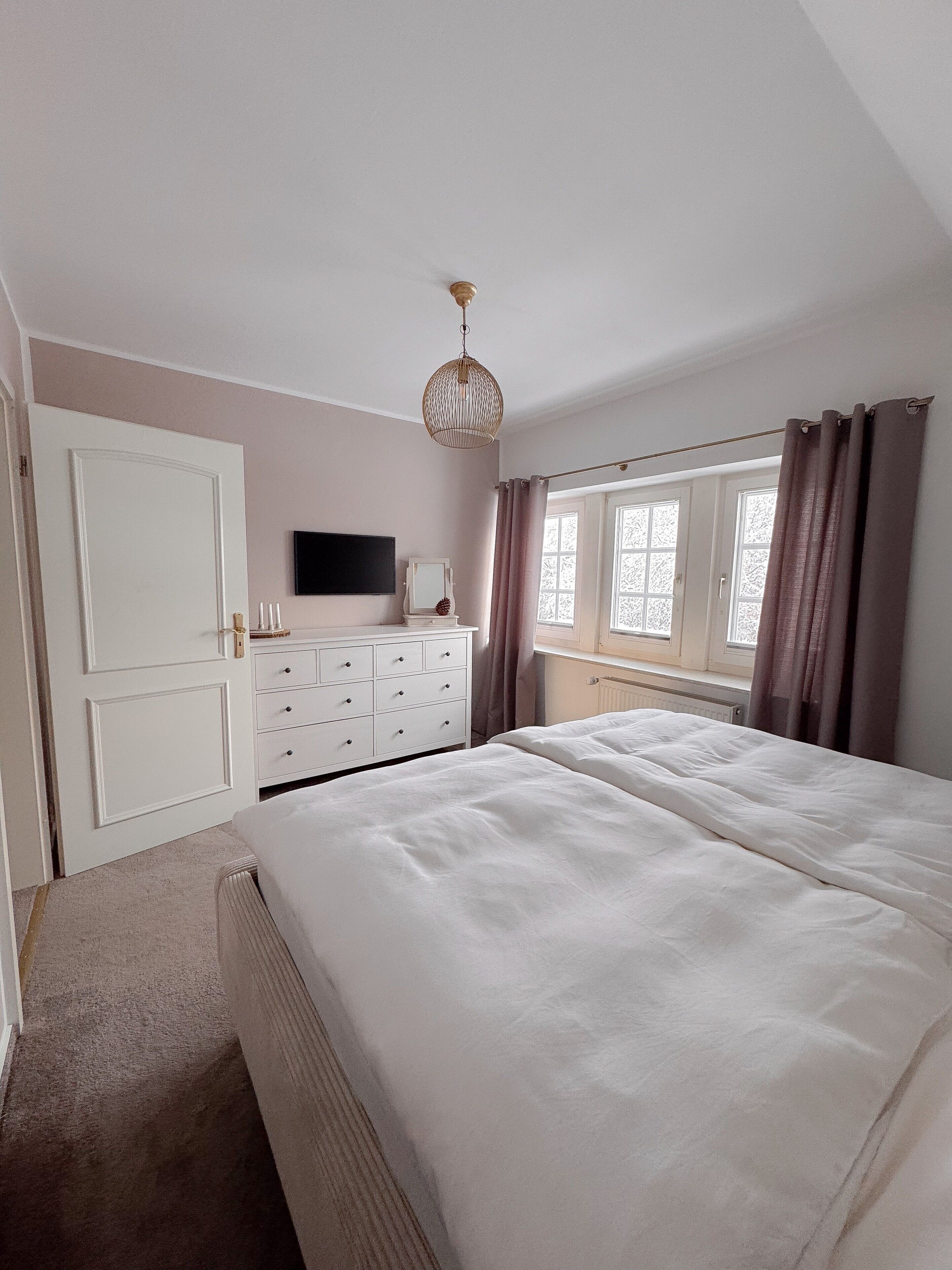Une chambre à coucher avec un lit, une commode, un téléviseur et une fenêtre avec des rideaux.