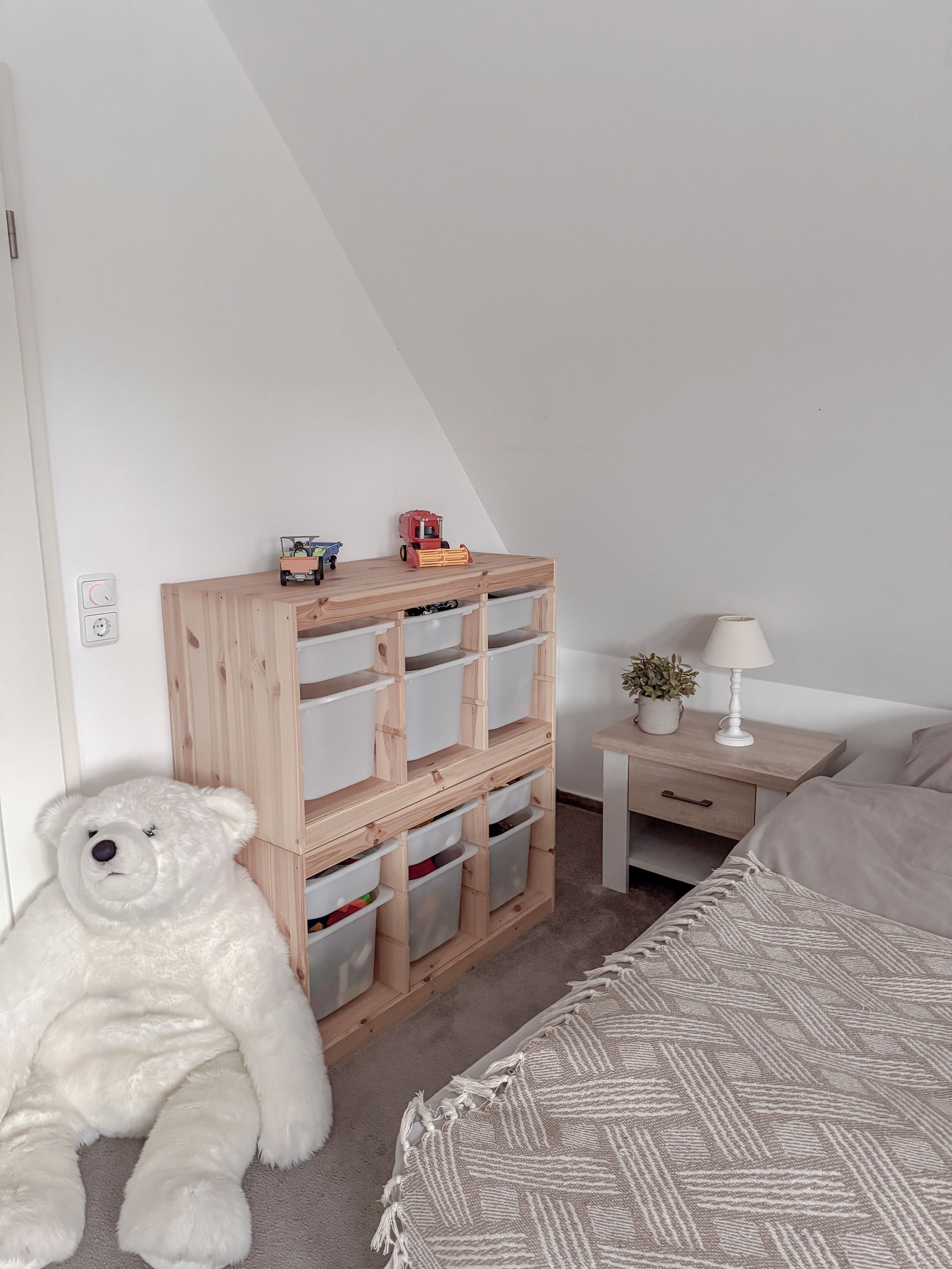 A bedroom with a bed, a wooden storage unit with drawers, a white teddy bear, a lamp, and a small plant.