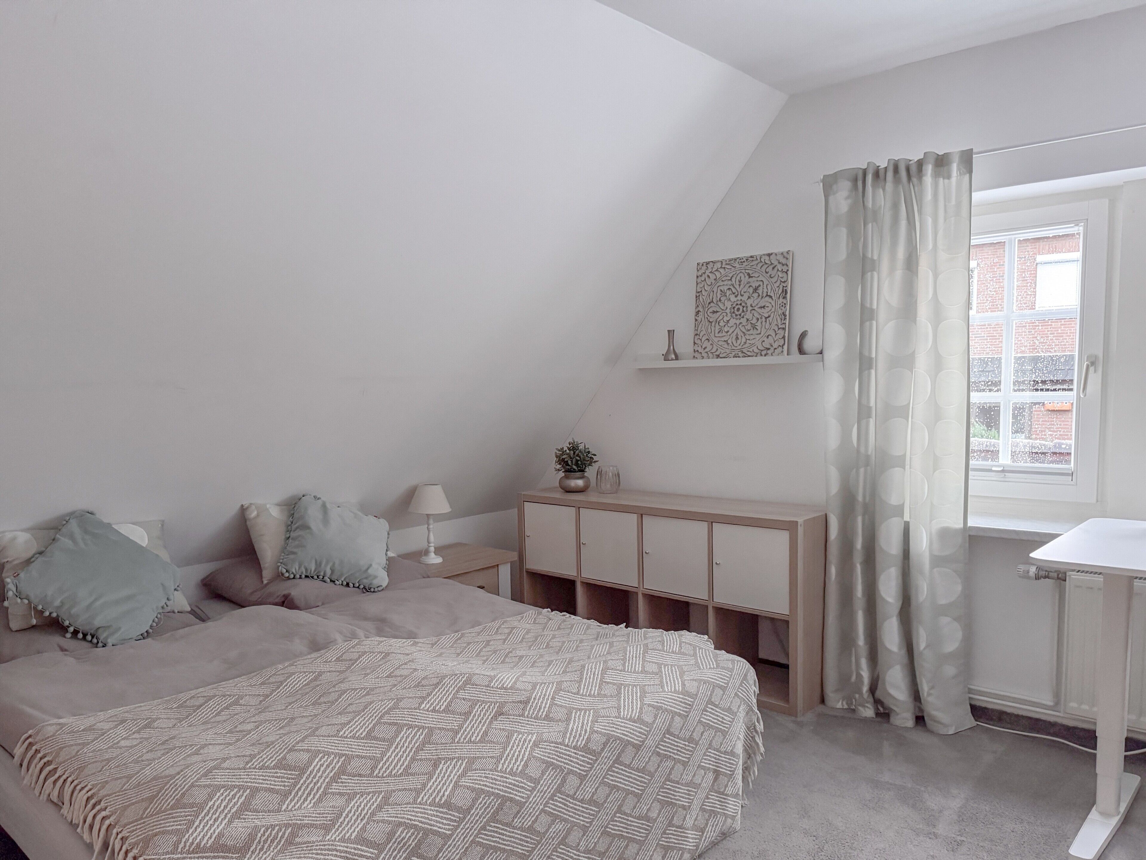 Een slaapkamer met een schuine wand, een bed met een bedovertrek met patroon, een nachtkastje met een lamp, een kast met geopende deuren en een raam met gordijn.
