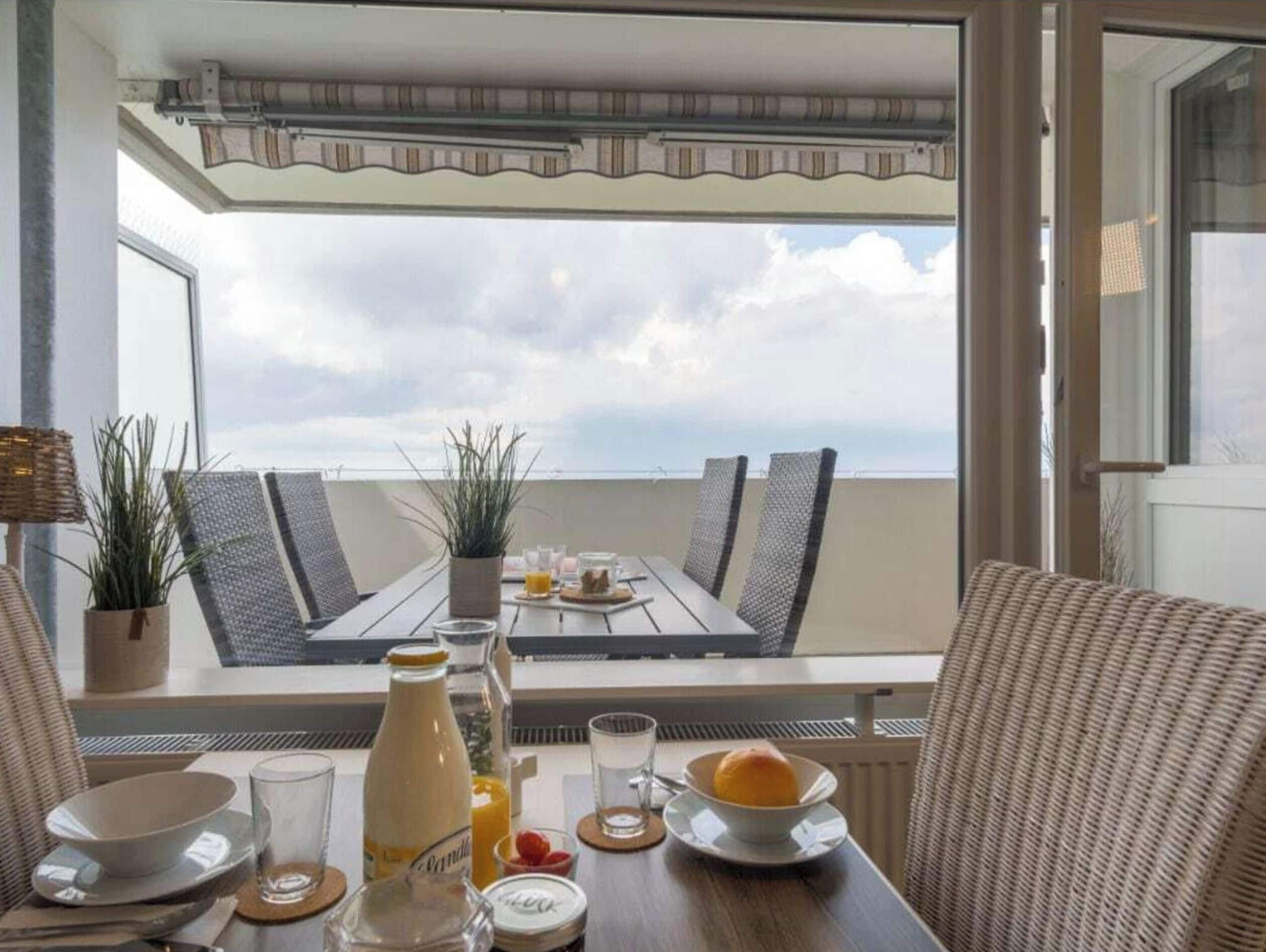 A dining table with chairs, a bottle, glasses, and a bowl of fruit on a balcony overlooking the sea.