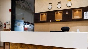 Balcão de recepção com painel frontal semelhante a mármore, relógios acima e um extintor de incêndio à esquerda.