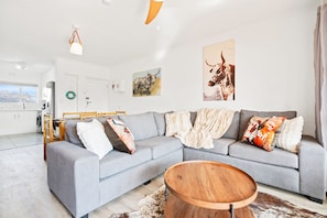 A modern living room with a grey sofa, a wooden coffee table, and a kitchen in the background. - Annies Haven at Greenways Golf Estate (Cape Town)