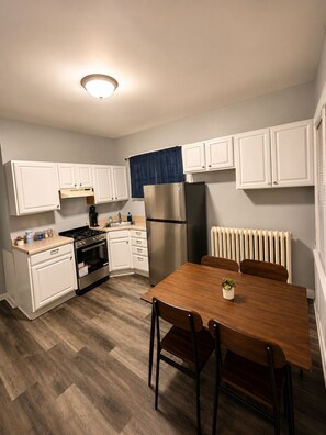 A compact kitchen with white cabinets, a stainless steel refrigerator, a wooden dining table with chairs, and a built-in oven.