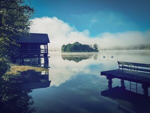 A tranquil lake with a wooden dock and a small wooden structure, surrounded by trees and a misty island.
