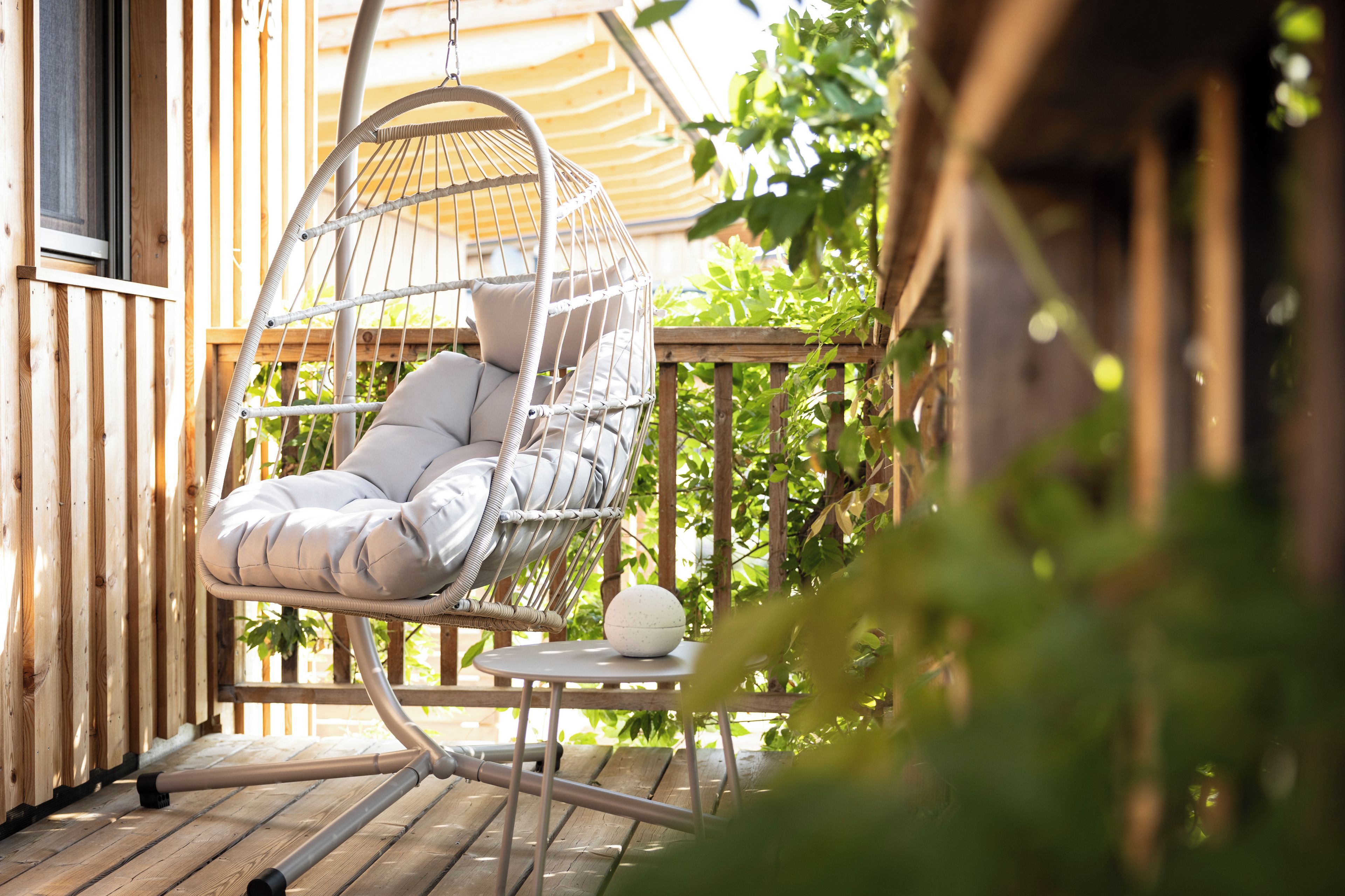 A wicker hanging chair with cushions on a wooden deck, a small table with a white spherical object, and a wooden fence with greenery in the background.