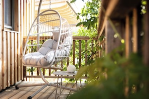 A wicker hanging chair with cushions on a wooden deck, a small table with a white spherical object, and a wooden fence with greenery in the background.