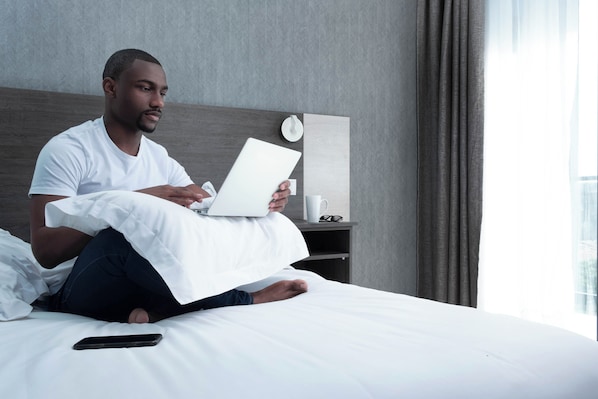 A person sitting on a bed with a laptop, a remote control, and a smartphone on the bed. There is a bedside table with a cup and a clock. - Elegant Beach Villa – Spacious, Private & Relaxing (Maputo)