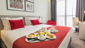 Hotellihuone, jossa on suuri sänky, punavalkoiset vuodevaatteet, aamiaistarjotin, jossa on munia, paahtoleipää ja mehua, sekä työpöytä ja tuoli.