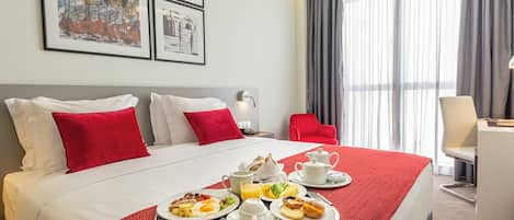 A hotel room with a large bed, red and white bedding, a breakfast tray with eggs, toast, and juice, and a desk with a chair.